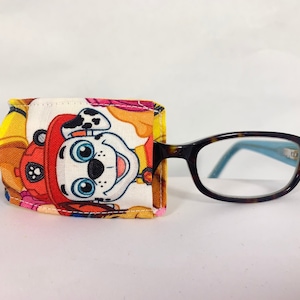 May include: A colorful fabric eyeglass holder with a cartoon dog character, featuring a red hat and white and black spots, holding a blue bone. The holder is attached to a pair of brown tortoise shell eyeglasses.