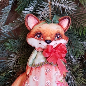 May include: A handmade felt fox ornament wearing a pink dress with white flowers and a red bow. The fox has a green bow on its head and is holding a red bow in its paws. The ornament is hanging from a green pine branch.