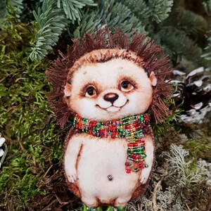 May include: A handmade hedgehog ornament with a brown fuzzy body, a white face with brown eyes, and a colorful beaded scarf. The ornament is wearing green shoes and is sitting on a bed of moss.