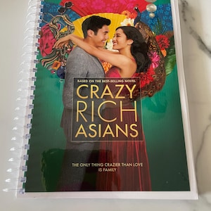 May include: A book cover for the movie "Crazy Rich Asians" based on the best-selling novel. The cover features a couple in front of a colorful background with peacocks and flowers. The text on the cover reads "Based on the best-selling novel Crazy Rich Asians The only thing crazier than love is family."