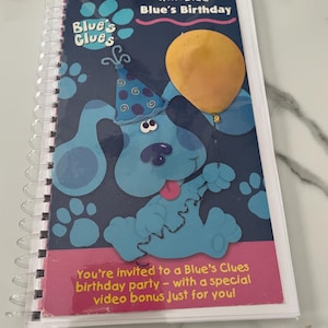 May include: A blue spiral bound book with a blue dog character on the cover. The dog is wearing a party hat and holding a yellow balloon. The book title is "Play Along With Blue - Blue's Birthday". The text "You're invited to a Blue's Clues birthday party - with a special video bonus just for you!" is on the bottom of the cover.