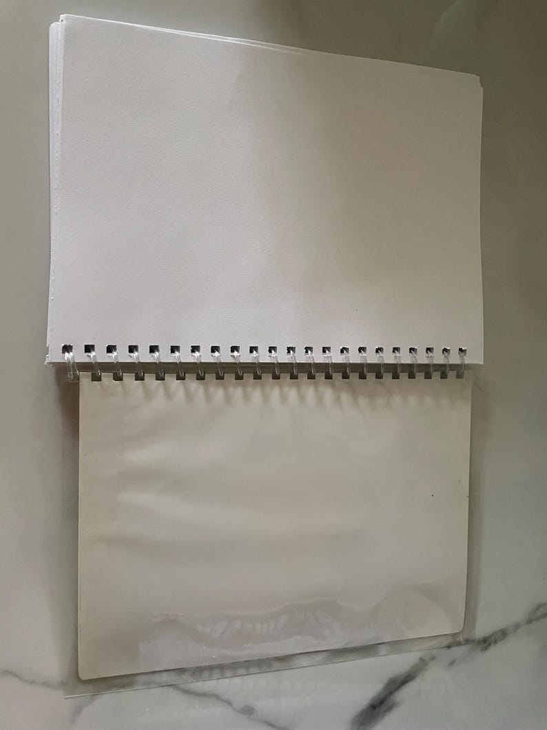May include: A white spiral bound notebook with a clear plastic cover. The notebook is blank and ready to be used for writing, drawing, or sketching.