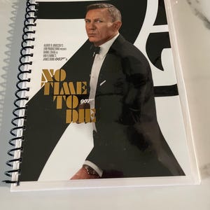 May include: A spiral-bound notebook featuring the movie poster for "No Time to Die." The cover shows Daniel Craig in a tuxedo, with the film's title in gold lettering. The notebook is labeled "2-DISC COLLECTOR'S EDITION."