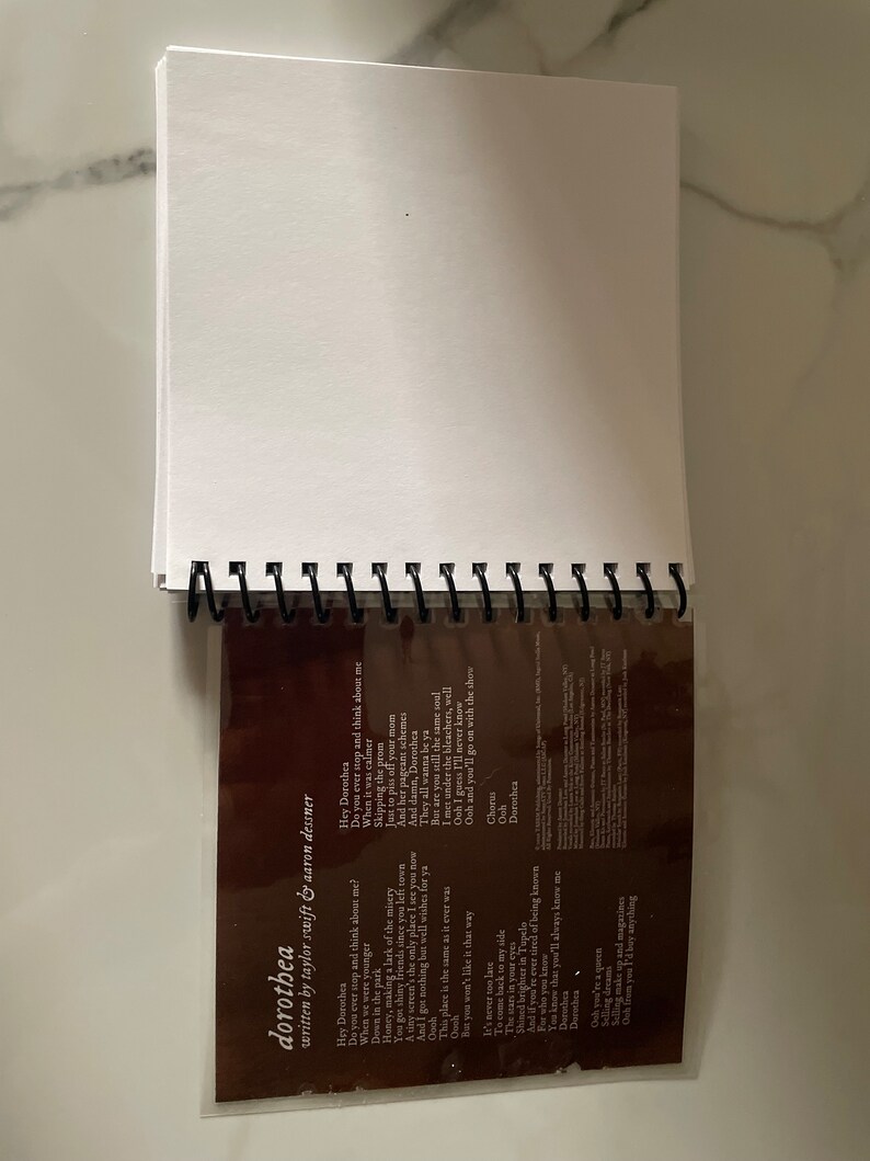 May include: A close-up of a brown notebook with a plastic sleeve covering the lyrics to the song "Dorothea" by Taylor Swift. The lyrics are printed in white text and include the lines "Hey Dorothea, Do you ever stop and think about me?" and "This place is the same as it ever was."