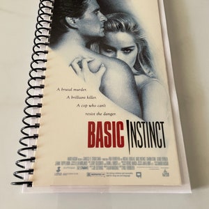 May include: A spiral bound notebook with a movie poster cover featuring Michael Douglas and Sharon Stone in the film "Basic Instinct". The poster has the text "A brutal murder. A brilliant killer. A cop who can't resist the danger." and the title "Basic Instinct".
