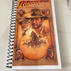 May include: Spiral-bound notebook featuring the movie poster for "Indiana Jones and the Last Crusade." The cover art showcases Harrison Ford as Indiana Jones, with other characters and the film's title prominently displayed. The notebook has a white border.
