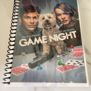 May include: A spiral-bound notebook featuring the movie poster for "Game Night." The poster shows Jason Bateman, Rachel McAdams, and a dog. Game pieces and playing cards are visible on the table. The notebook has a clear plastic cover.