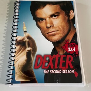 May include: A spiral-bound DVD case for "Dexter: The Second Season" featuring a portrait of the main character. The cover includes the show's title in red, the season title, and an image of a syringe. The case is encased in a clear plastic sleeve.