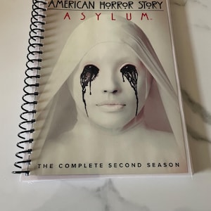 May include: A white spiral bound notebook with a black and white image of a woman with black tears running down her face. The text on the cover reads "American Horror Story Asylum: The Complete Second Season".