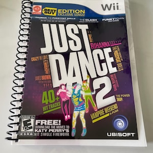 May include: A Just Dance 2 Wii game case with a spiral binding. The cover features the game title in large white letters, with colorful dancer figures and song titles. Includes the text "40+ Hit Tracks" and "Free! Download the moves to Katy Perry's Hit Single Firework".