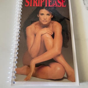 May include: A spiral bound notebook with a cover featuring a photo of Demi Moore from the movie "Striptease". The cover is black with the title "Striptease" in red letters and Demi Moore's name in white letters.