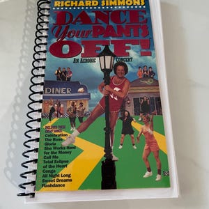 May include: A spiral-bound book titled "DANCE YOUR PANTS OFF!" by Richard Simmons. The cover features Simmons in a red outfit, posing by a lamppost. The book lists songs like "Celebration" and "Flashdance".
