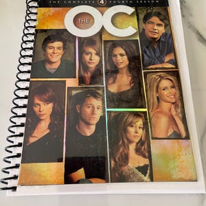 Puede incluir: Un set de DVD encuadernado en espiral de "The OC: The Complete Fourth Season". La portada presenta un collage de retratos de personajes sobre un fondo degradado dorado y naranja. El título del programa se muestra de forma destacada.