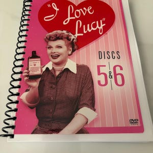 May include: A spiral-bound DVD case featuring a pink and white striped background with a large red heart containing the text I Love Lucy. The cover displays a photograph of a woman holding a bottle, with the text Discs 5 & 6 printed in black.