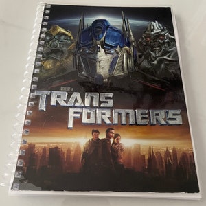May include: A black and silver spiral bound notebook with a Transformers movie poster on the cover. The poster features Optimus Prime, Bumblebee, and Megatron. The title "Transformers" is written in silver letters.