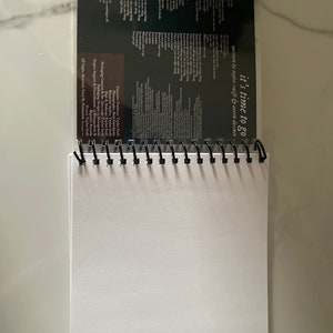 May include: A black spiral bound notebook with a clear plastic cover. The cover features a white text overlay that reads "it's time to go" and "written by taylor swift & aaron dessner".
