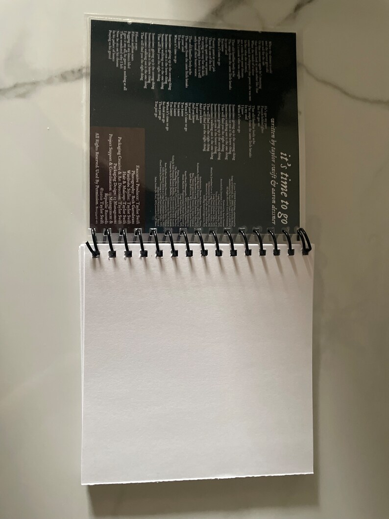 May include: A black spiral bound notebook with a clear plastic cover. The cover features a white text overlay that reads "it's time to go" and "written by taylor swift & aaron dessner".