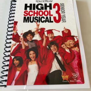 Pode incluir: Um caderno de espiral com a capa do filme High School Musical 3: Senior Year. A capa mostra um grupo de jovens com becas e capelos de formatura vermelhos, com o título do filme em letras pretas e vermelhas em negrito sobre fundo branco.