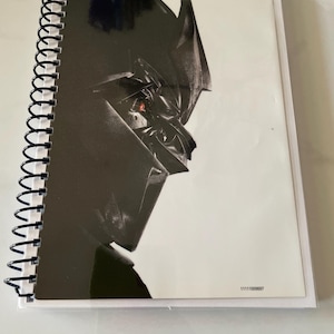May include: A black and white notebook with a spiral binding. The cover features a close-up of a robot's face with a red eye. The number 11111000097 is printed on the bottom right corner of the cover.