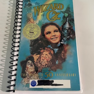 May include: A spiral-bound booklet featuring the movie "The Wizard of Oz." The cover has a blue background with images of characters like Dorothy, the Tin Man, and the Cowardly Lion. Text includes "Limited Edition" and "50th Anniversary."