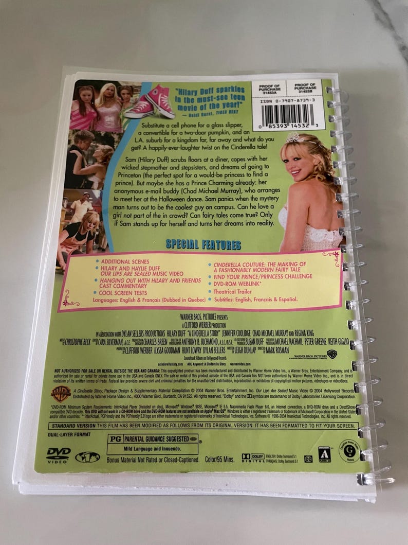 May include: DVD case for the movie "A Cinderella Story" starring Hilary Duff. The case is pink and green with a picture of Hilary Duff on the front. The back of the case lists special features, including additional scenes, music videos, and a behind-the-scenes look at the making of the movie.