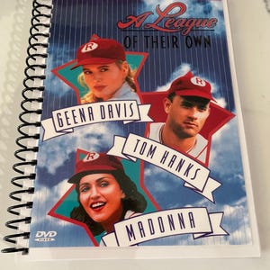 Puede incluir: Una funda de DVD con espiral para la película "A League of Their Own". La portada presenta imágenes de Geena Davis, Tom Hanks y Madonna, con el título de la película en una fuente estilizada. La funda está etiquetada como "Deluxe Widescreen Presentation".