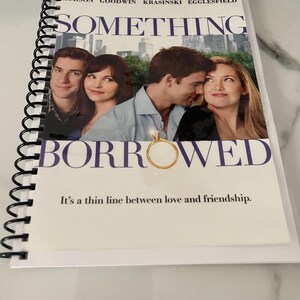May include: A spiral-bound notebook featuring the movie poster for "Something Borrowed." The poster shows the film's title and cast, with a romantic image of the actors. The tagline "It's a thin line between love and friendship" is also visible.