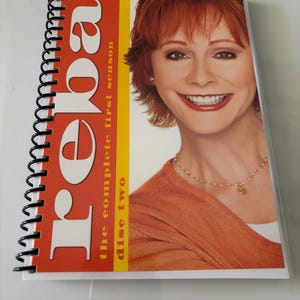 May include: A spiral-bound DVD case for Reba: The Complete First Season, Disc Two. The cover features a portrait of a person with reddish-brown hair wearing an orange sweater and a gold necklace, with the text Reba, the complete first season, disc two.