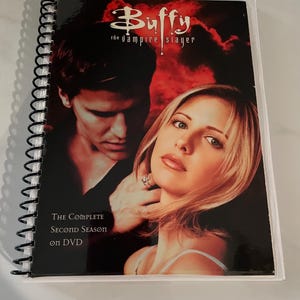Könnte beinhalten: Eine spiralgebundene DVD-Hülle für "Buffy the Vampire Slayer: Die komplette zweite Staffel". Das Cover zeigt einen dunklen Hintergrund mit Bildern von zwei Personen und dem Logo der Show. Der Text auf dem Cover lautet "Die komplette zweite Staffel auf DVD."