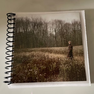 May include: A person wearing a plaid coat walks through a field of tall grass and trees in the background. The photo is taken on a cloudy day.