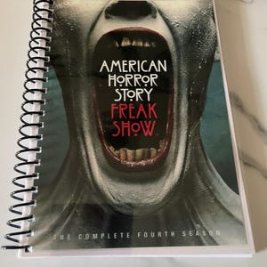 May include: A spiral bound notebook with a cover featuring a close-up of a person's open mouth with a red and white mouth and teeth. The text "American Horror Story Freak Show" is printed in white on a black background. The text "The Complete Fourth Season" is printed in white on a black background at the bottom of the cover.