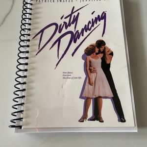 Może przedstawiać: Biały notes ze spiralą i okładką z fotografią Patricka Swayze i Jennifer Grey z filmu "Dirty Dancing". Tytuł filmu jest napisany fioletowym pismem na górze okładki. Tekst "First dance. First love. The time of your life." jest wydrukowany pod zdjęciem.