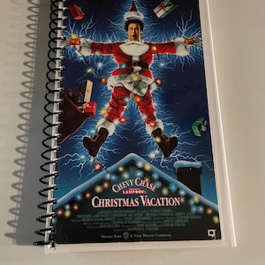 May include: A spiral-bound notebook featuring the movie poster for "National Lampoon's Christmas Vacation." The poster shows a man in a Santa suit with the text "Yule Crack Up!" at the top. The notebook is white with a black spiral binding.