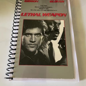 May include: A spiral-bound notebook featuring the movie poster for "Lethal Weapon." The cover shows Mel Gibson and Danny Glover, with text including "Special Widescreen Edition" and the film's title. The notebook is encased in a clear plastic sleeve.