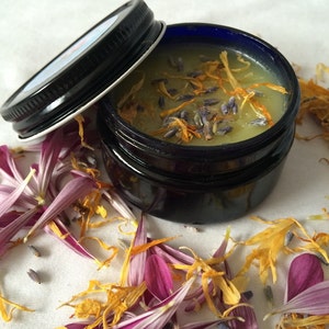 May include: A small glass jar with a black lid filled with a light green balm. The balm is topped with dried lavender and yellow flower petals.