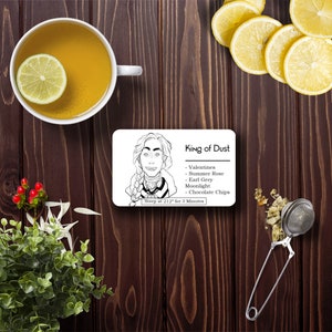 May include: A top-down view of a cup of tea with a lemon slice, a card labelled "King of Dust" with a drawing, and a tea infuser on a dark wooden surface. The card lists tea flavours.