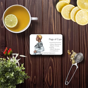 Puede incluir: Una tarjeta blanca con las palabras "Page of Cups" y una lista de mezclas de té: White Blueberry, Maple Cream Oolong, White Monkey, Cornflowers. La tarjeta también incluye instrucciones para preparar el té a 195 grados durante 3 minutos.