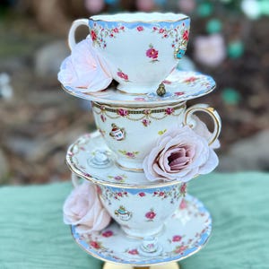 May include: Stacked vintage teacups and saucers with floral patterns and gold trim, adorned with pink roses. The teacups are white with blue rims and delicate floral designs. The base is a gold-colored stand.