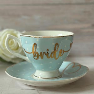 May include: A light blue teacup and saucer set with gold polka dots and the word "bride" written in gold script.