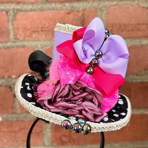 May include: A miniature top hat headband with a black and white polka dot brim, adorned with pink, purple, and fuchsia ribbons, sequins, and a cluster of iridescent beads. The hat is attached to a black headband.