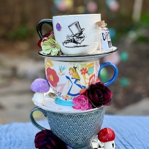 May include: Stacked teacups with Alice in Wonderland theme. The top cup features a Mad Hatter illustration and a small bottle with "DRINK" text. The middle cup has an Alice illustration, and the bottom cup is gray with a geometric pattern. Decorative flowers and mushrooms are included.
