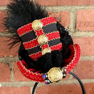 May include: A black and red miniature top hat headband featuring gold-toned buttons, red decorative trim, black feathers, and iridescent beads. The hat is adorned with three vertical gold buttons on the front and one centered on the brim.