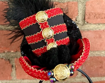 Peut inclure: Un serre-tête avec un chapeau haut-de-forme miniature noir et rouge, orné de boutons dorés, de galons rouges, de plumes noires et de perles irisées. Le chapeau présente trois boutons dorés verticaux et un bouton central sur le bord.