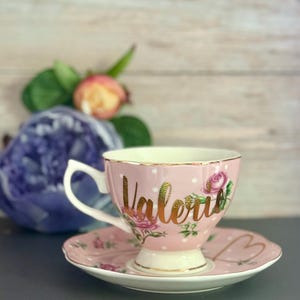 May include: A pink teacup and saucer set with gold trim and the name "Valerie" in gold script. The cup and saucer are decorated with white polka dots and floral designs. The saucer has a heart shape.