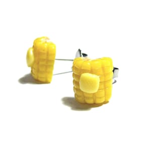 Puede incluir: Un par de pendientes de maíz amarillo con un pequeño trozo de mantequilla en la parte superior. Los pendientes están hechos de arcilla polimérica y tienen ganchos de plata.