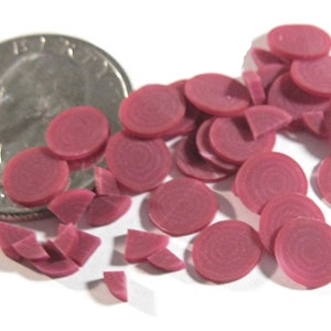 May include: A collection of small, round, dark pink discs and several quarter-circle pieces. The discs have a concentric circle pattern. The objects are scattered on a white surface next to a US quarter coin.