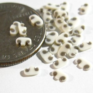 Puede incluir: Una colección de pequeños amuletos de cerámica blanca en forma de hongo con manchas marrones. Los amuletos están esparcidos sobre una superficie blanca junto a una moneda de veinticinco centavos de dólar estadounidense.