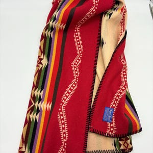 May include: Red and cream colored woven blanket with a geometric pattern. The blanket has a black border and a blue label that says "Pendleton."