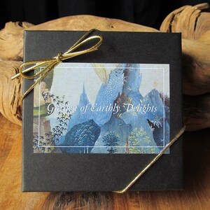 May include: A black gift box with a gold ribbon tied around it. The box has a white label with the text "Garden of Earthly Delights" printed on it. The label features a blue and white illustration.