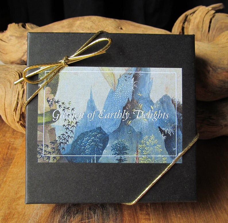 May include: A black gift box with a gold ribbon tied around it. The box has a white label with the text "Garden of Earthly Delights" printed on it. The label features a blue and white illustration.
